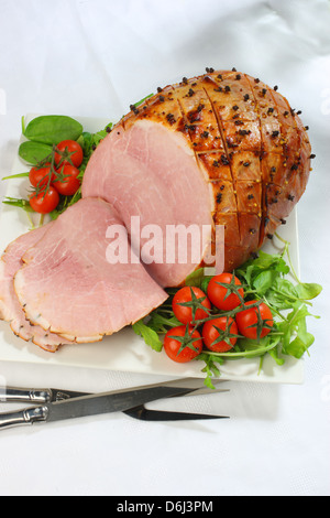 Jambon cuit au four avec des clous de girofle et les tomates cerises et couteau et fourchette sur un carré blanc sur la plaque d'une nappe blanche Banque D'Images