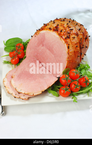 Jambon cuit au four avec des clous de girofle et les tomates cerises et couteau et fourchette sur un carré blanc sur la plaque d'une nappe blanche Banque D'Images