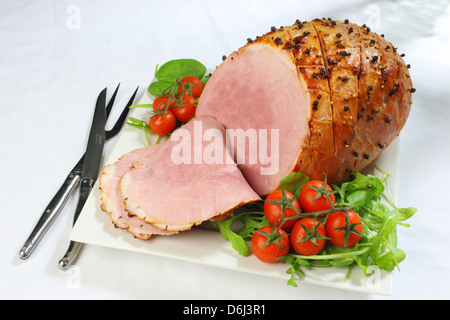 Jambon cuit au four avec des clous de girofle et les tomates cerises et couteau et fourchette sur un carré blanc sur la plaque d'une nappe blanche Banque D'Images