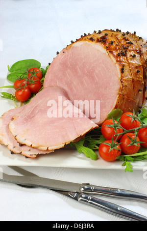 Jambon cuit au four avec des clous de girofle et les tomates cerises et couteau et fourchette sur un carré blanc sur la plaque d'une nappe blanche Banque D'Images