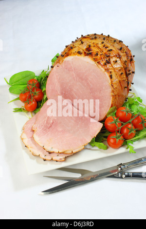 Jambon cuit au four avec des clous de girofle et les tomates cerises et couteau et fourchette sur un carré blanc sur la plaque d'une nappe blanche Banque D'Images