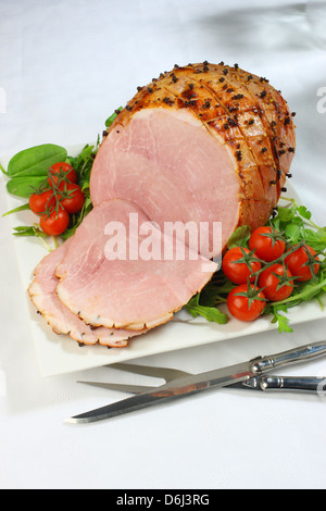 Jambon cuit au four avec des clous de girofle et les tomates cerises et couteau et fourchette sur un carré blanc sur la plaque d'une nappe blanche Banque D'Images