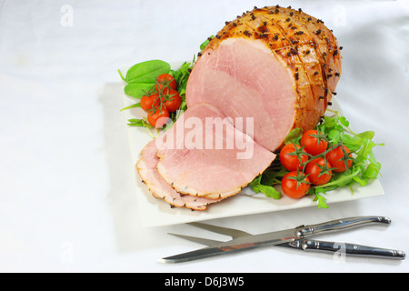 Jambon cuit au four avec des clous de girofle et les tomates cerises et couteau et fourchette sur un carré blanc sur la plaque d'une nappe blanche Banque D'Images