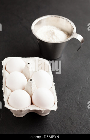 Les oeufs de poule en carton blanc avec la farine dans une tasse à mesurer en métal sur fond sombre Banque D'Images