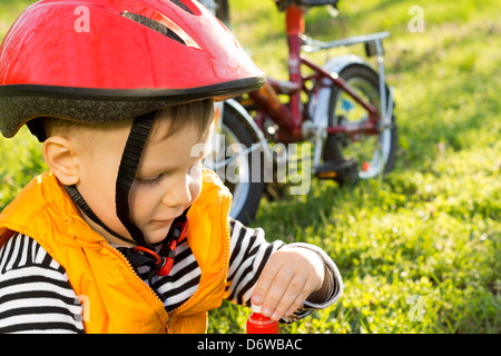 Petit garçon à vélo assis sur l'herbe verte de boire de l'eau en bouteille avec sa moto garée à côté et portant un casque de sécurité Banque D'Images