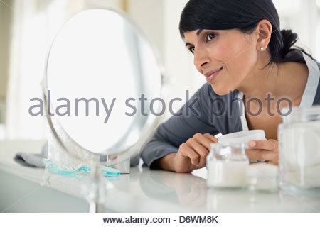 Belle femme tenant une bouteille de crème tout en regardant dans le miroir de coiffeuse