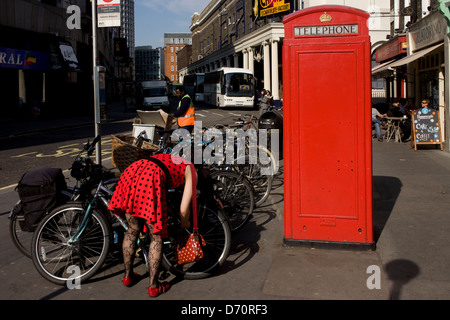 Une dame portant une robe rouge à pois noir verrouille son vélo près d'une cabine téléphonique publique rouge. Banque D'Images