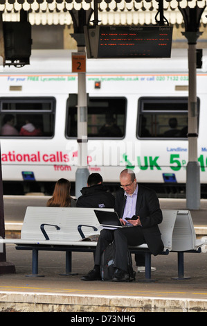 Un homme d'affaires assis sur une gare ferroviaire de la plate-forme à l'aide d'un ordinateur portable tout en attendant son train Banque D'Images