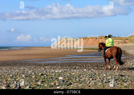 Cavalière à cheval sur une plage de galets à marée basse contre un ciel bleu avec des nuages blancs Banque D'Images