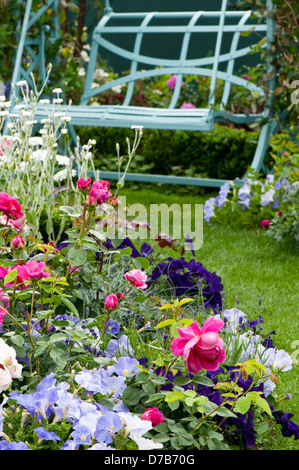 Image Portrait d'un jardin en été, avec de nombreuses fleurs colorées bordant une pelouse menant à un siège en fer. Banque D'Images