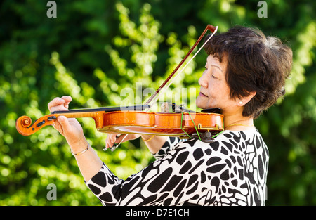 Photo horizontale de Senior Asian woman facing latéralement tout en jouant du violon vert en plein air avec des arbres en arrière-plan Banque D'Images