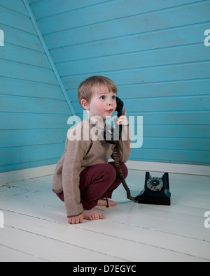 Un garçon jouant avec un vieux téléphone à cadran rotatif assis sur le plancher en bois blanc, pieds nus dans une chambre loft bleu Banque D'Images