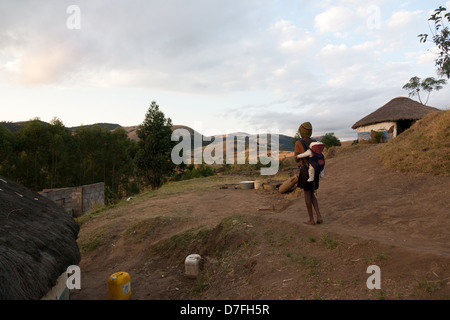 Une jeune femme africaine de marcher dans les collines près de sa maison, portant un bébé sur son dos Banque D'Images