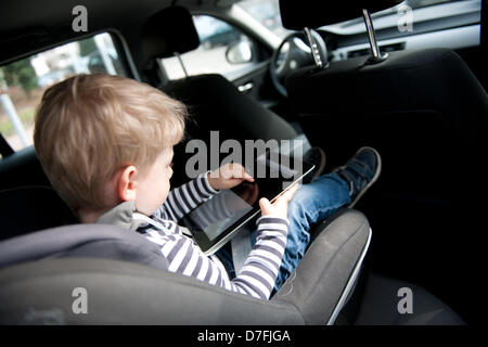 Un garçon est assis dans son siège pour enfants dans une voiture à jouer avec un ipad. Banque D'Images