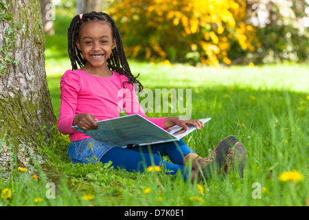 Outdoor portrait of a cute young black petite fille lisant un livre - les peuples africains Banque D'Images