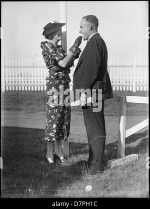 Cette photographie en noir et blanc montre un couple à l'hippodrome de Warwick Farm en Australie, profitant d'un moment en plein air. La femme tient de la crème glacée tandis que l'homme se tient à côté d'elle, portant un chapeau et des gants. L'image capture la mode et l'atmosphère d'une sortie sur l'hippodrome, reflétant le style du milieu du XXe siècle et les activités de loisirs associées à cet endroit australien. Banque D'Images