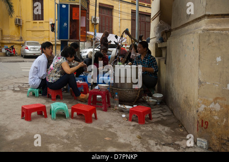 Vue horizontale d'un étal de vente de nouilles Pho, traditionnels, à des gens heureux de s'asseoir sur un tabouret en plastique sur le trottoir. Banque D'Images