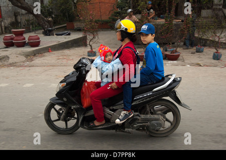 L'horizontale portrait d'une femme montant un cyclomoteur avec son bébé dans un faisceau et son fils équitation leurs à Hanoi. Banque D'Images