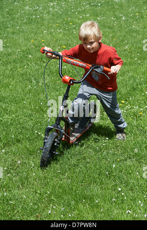 Un jeune enfant garçon blond sur un scooter, parc d'été Banque D'Images