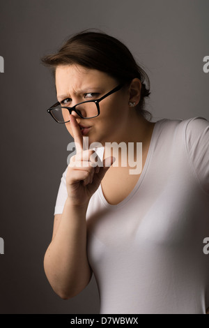 Une jeune brunette young woman wearing glasses avec son doigt sur ses lèvres, demandant le silence Banque D'Images