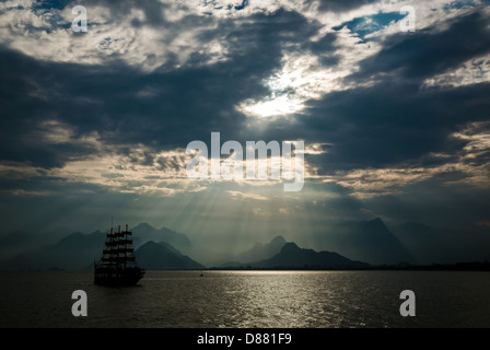 Bateau à voile à traverser la mer en face de l'horizon la montagne sous le ciel nuageux. Les rayons du soleil Banque D'Images