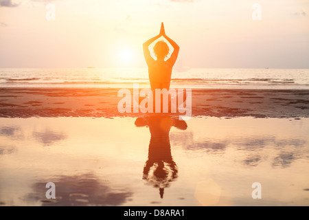 Yoga woman sitting in lotus pose sur la plage pendant le coucher du soleil, avec la réflexion dans l'eau (en couleurs)
