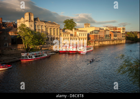 Une flotte de bateaux de croisière et de bateaux touristiques amarrés le long du Guildhall lors d'une soirée d'été ensoleillée avec un bateau à rames au premier plan.River Ouse, York. Banque D'Images