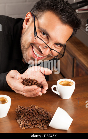 Un homme aux cheveux foncés, à la barbe et aux lunettes est penché sur une table en bois, souriant à la caméra. Il tient une poignée de grains de café torréfiés dans ses mains gobetées. Sur la table, il y a deux blancs Banque D'Images