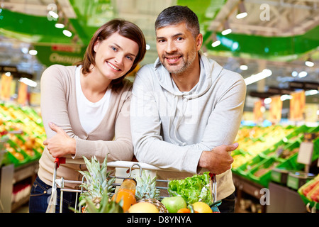 Image de l'heureux couple avec panier remplis de produits looking at camera in supermarket Banque D'Images