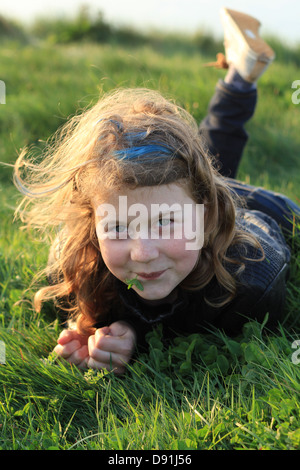 Jeune fille de 12 ans souriante portant sur l'herbe avec le trèfle dans sa bouche et le coucher du soleil la lumière dans ses cheveux blonds Banque D'Images