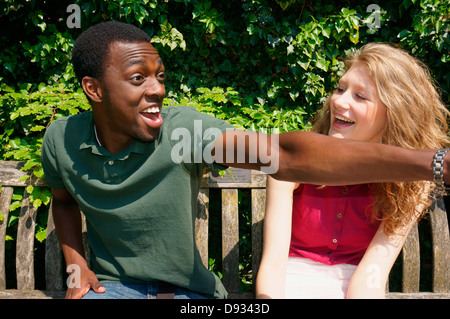 Couple heureux, assis sur un banc à l'extérieur, au soleil, avec un petit ami excité du doigt quelque chose, à l'amusement de sa petite amie. Banque D'Images