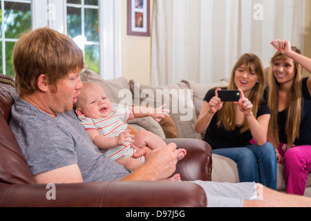 Père holding baby boy alors que mère avec sa sœur essaient de prendre une photo sur un téléphone intelligent caméra dans la salle de séjour Banque D'Images