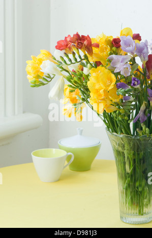 Une bande de jaune, rouge, violet, rose, et blanc Freesia dans un vase en verre sur un tapis de table jaune. Banque D'Images