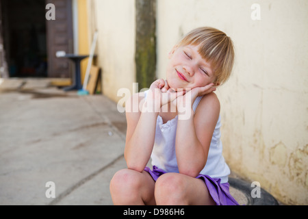 Portrait of cute smiling young girl with eyes closed assis en face de mur de béton à l'extérieur Banque D'Images