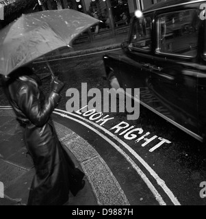 Un piéton est sur le point d'étape sur une rue du centre de Londres, en traversant les mots Regardez à droite comme un taxi tourne à gauche. Vu en noir et blanc, nous voyons des flocons gelés par flash sur ce coin de rue urbaine dans la ville de Londres, au cœur de la capitale, le quartier financier. Le stencil marcheurs mots racontent à regarder les voitures venant en sens inverse, lorsqu'il n'est pas facile de ne pas se concentrer et la cause de nombreuses blessures (principalement pour les touristes) à ceux qui ne sont pas utilisées pour le trafic sur le côté opposé de la route pour le reste de l'Europe. Banque D'Images