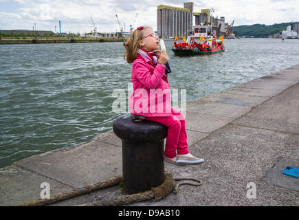 Petite fille de manger des glaces, assis sur un poteau à l'Armada de Rouen. Vue sur la rivière et l'arrière-plan industriel. Petite fille en premier plan le port de pantalon rouge et rose. Tugboat et silos en arrière-plan. Crédit : Christine Gates/Alamy Live News Banque D'Images