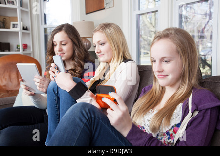 Girls sitting on sofa using digital tablet, d'un téléphone cellulaire et de jeu vidéo de poche Banque D'Images