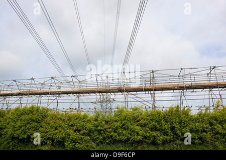 Échafaudage d'entretien sous les fils de l'électricité haute tension sous un ciel nuageux avec haie d'aubépine en premier plan. Banque D'Images