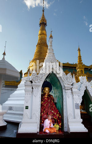 La Schwedagon, temple sacré de birmanie au coeur de Rangoon. Une religieuse dans sa robe rose mange devant un Bouddha dans une chapelle latérale. Banque D'Images