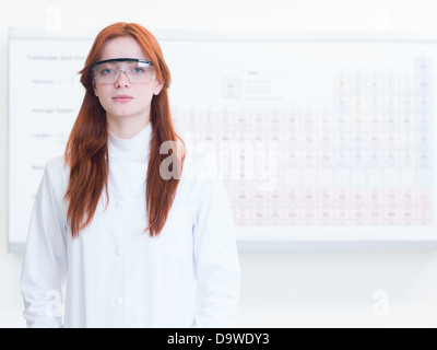 Close-up of attractive redhead caucasian girl habillés en uniforme d'un scientifique avec des lunettes de sécurité, avec tableau périodique en background Banque D'Images