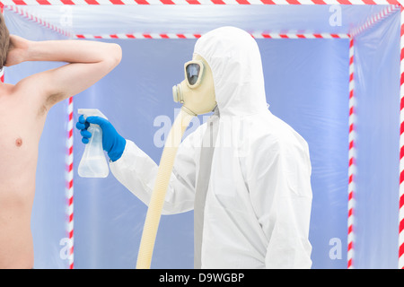 Personne dans biohazard costume et masque à gaz de la pulvérisation de désinfectant l'aisselle d'un homme portant pas de chemise en face d'une tente de confinement Banque D'Images