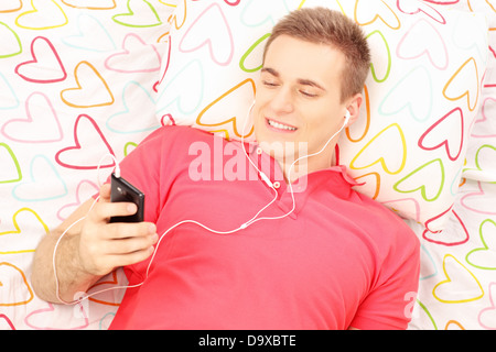 Young smiling man allongé sur un lit et l'écoute de la musique à partir de son téléphone Banque D'Images