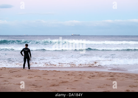 Un surfeur solitaire se dresse sur le sable doré en regardant les vagues sur une plage de surf à Wollongong, Australie. Banque D'Images