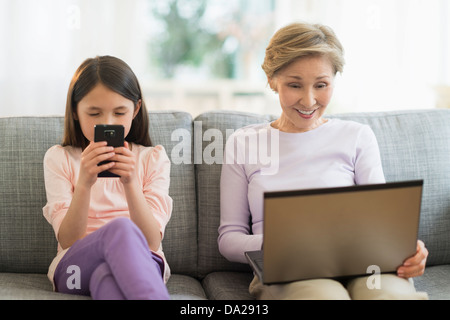 Petite-fille (8-9) et la grand-mère assise sur un canapé et à l'aide d'ordinateur portable et téléphone cellulaire Banque D'Images