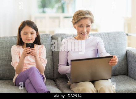 Petite-fille (8-9) et la grand-mère assise sur un canapé et à l'aide d'ordinateur portable et téléphone cellulaire Banque D'Images