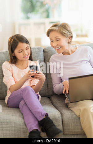 Petite-fille (8-9) et la grand-mère assise sur un canapé et à l'aide d'ordinateur portable et téléphone cellulaire Banque D'Images