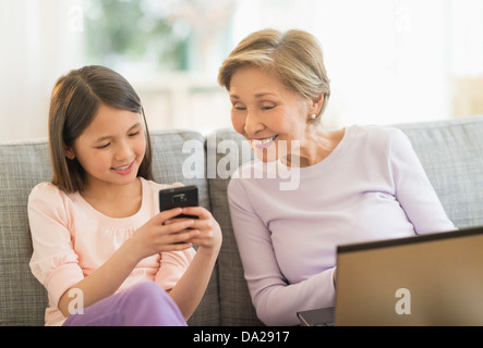 Petite-fille (8-9) et la grand-mère assise sur un canapé et à l'aide d'ordinateur portable et téléphone cellulaire Banque D'Images