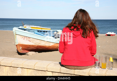 Woman in red jacket assis sur le mur face à la plage Banque D'Images