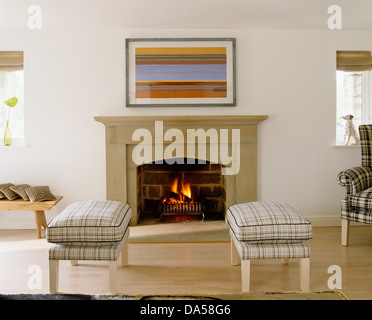 Paire de tabourets rembourrés cochée en face de simple cheminée avec feu allumé en pays moderne salon avec parquet en bois Banque D'Images
