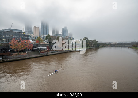 Un rameur exercices sur la rivière Yarra et l'horizon brumeux du centre-ville de Melbourne, Australie. Banque D'Images